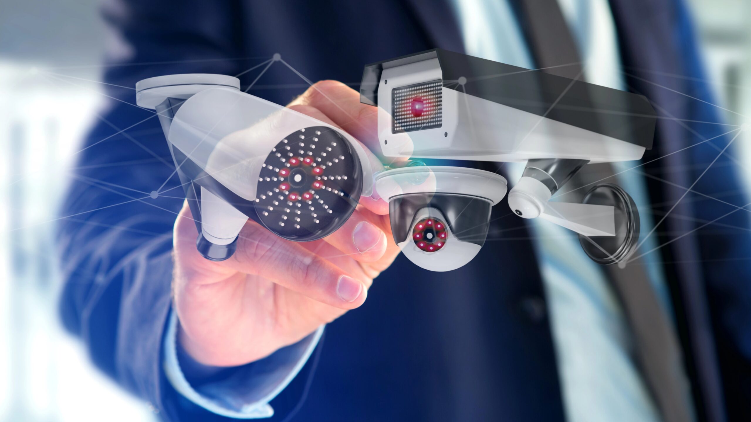 Das Bild symbolisiert die Videoüberwachung durch den Arbeitgeber, indem drei kleine Kameras gezeigt werden und ein Hand vor dem Torso eines Mannes im Hintergrund, wie dieser sich Notizen macht.